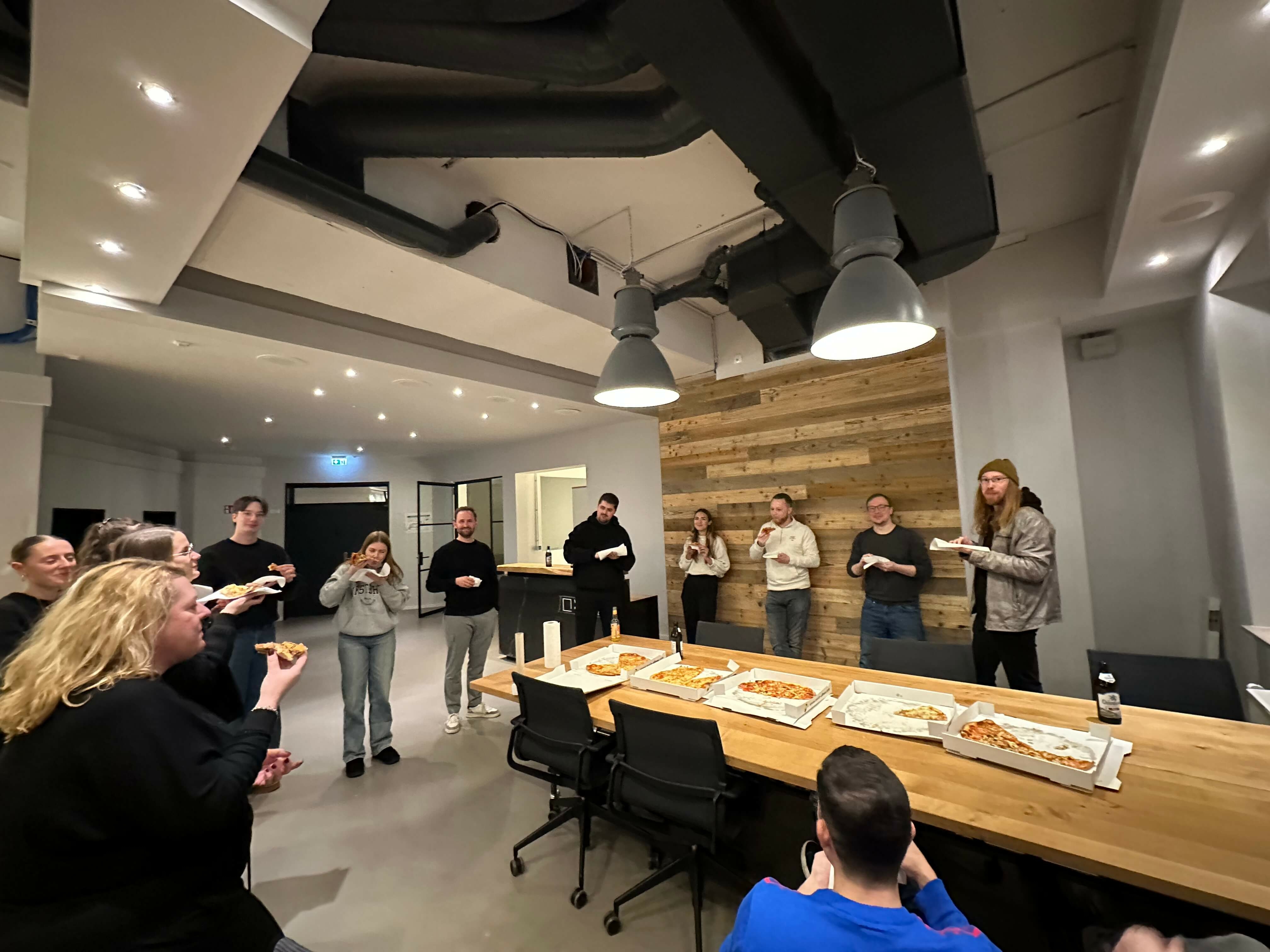 Gruppe von Personen in modernem Büro sitzt und steht um einen langen Holztisch und isst Pizza.