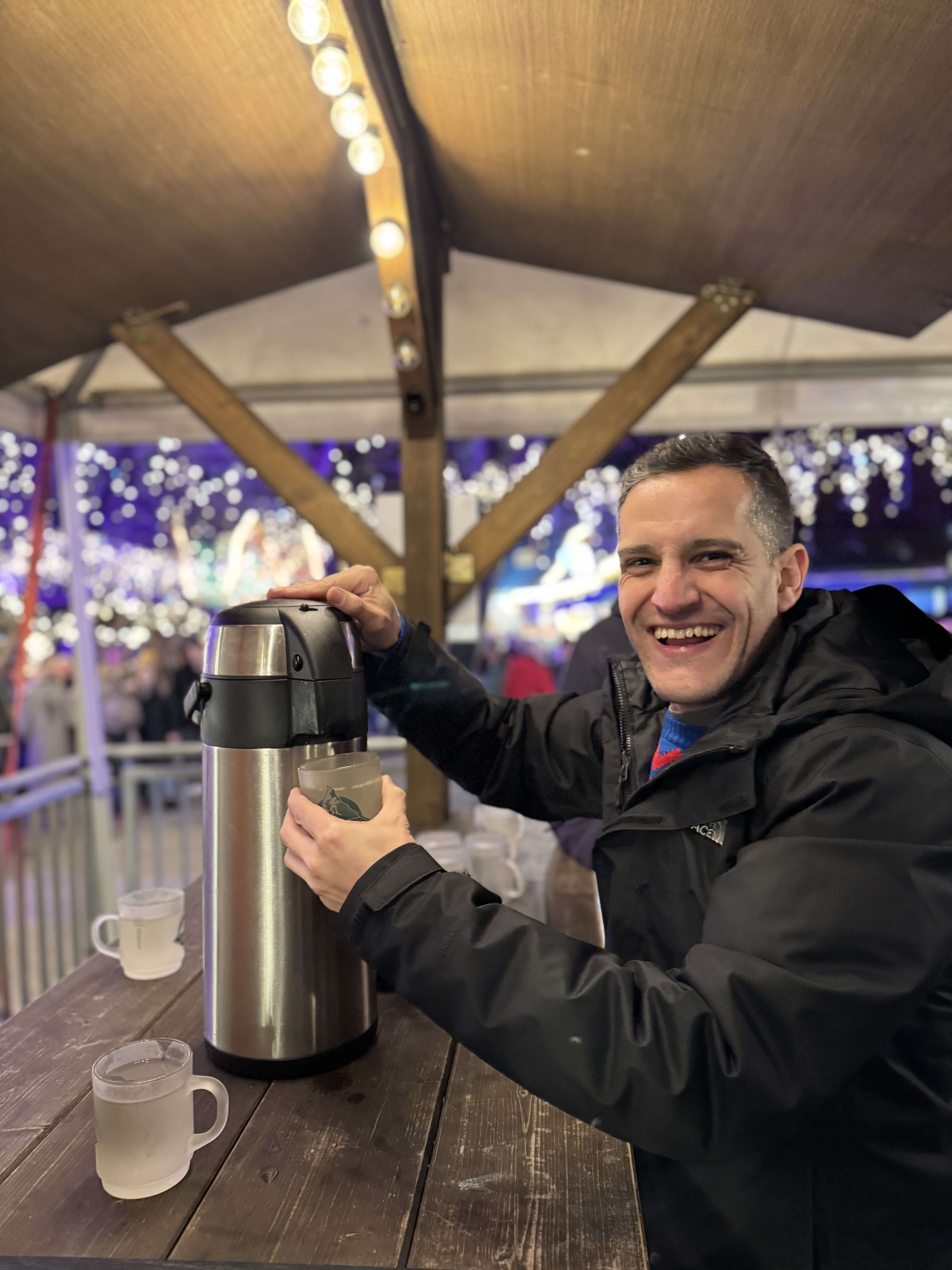 Mann in dunkler Jacke steht an einem Getränkestand und zapft sich ein Heißgetränk aus einer großen Thermoskanne; im Hintergrund Lichter und Stände auf einem Weihnachtsmarkt.