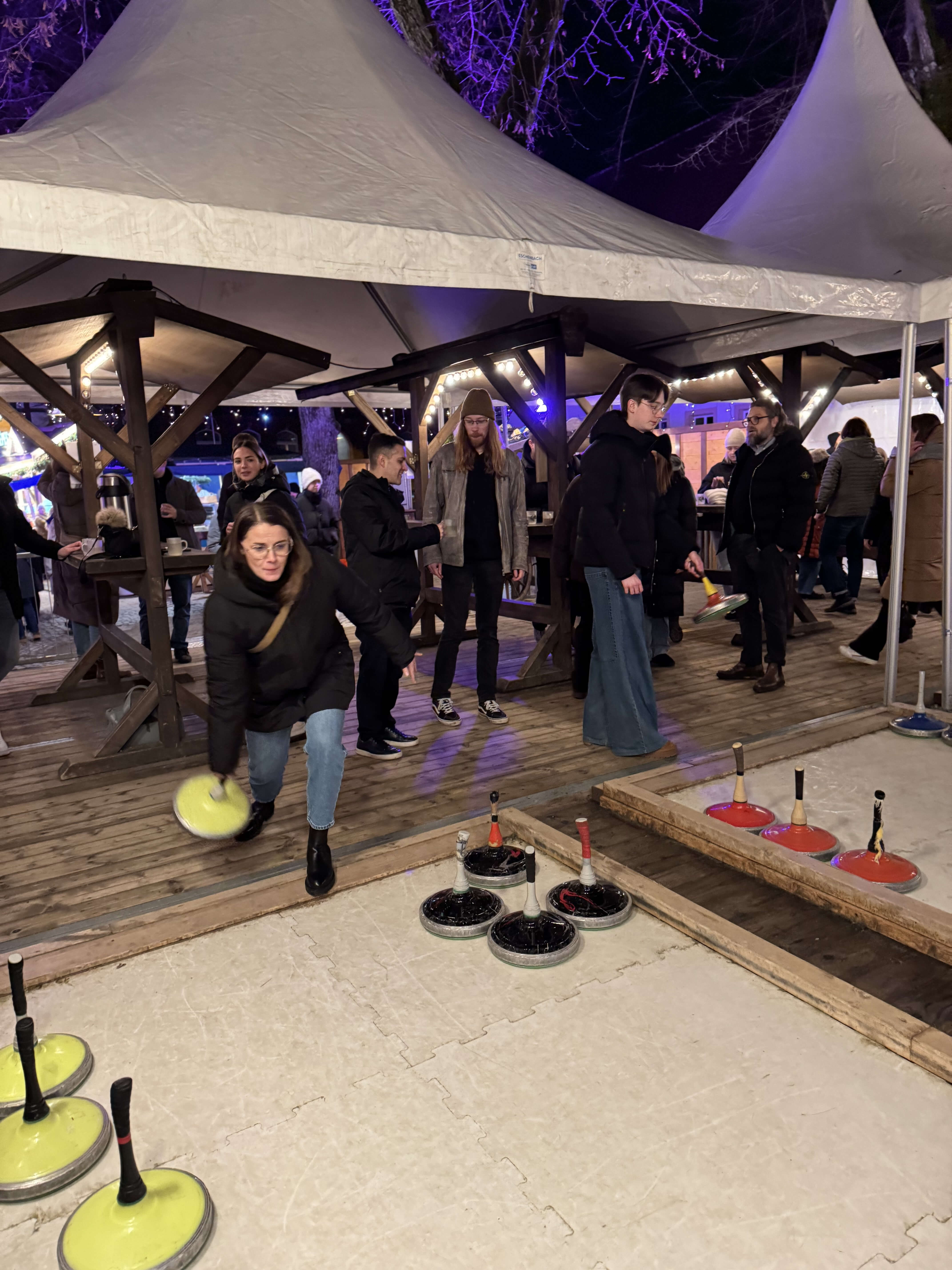 Menschen spielen abends draußen Eisstockschießen auf einer beleuchteten Bahn beim Weihnachtsmarkt.