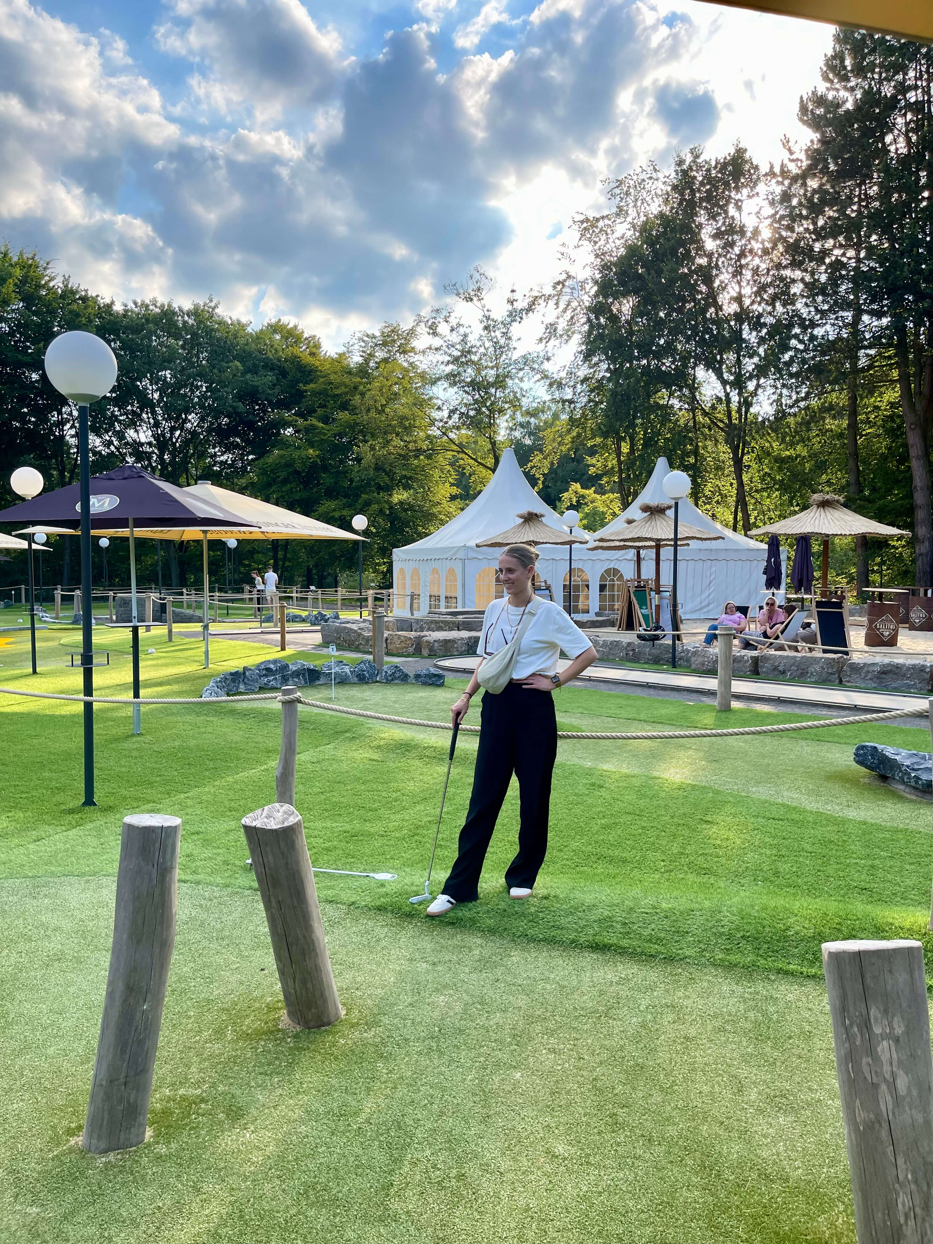Person steht auf einer Adventure-Golf-Bahn auf einer Grünfläche; im Vordergrund sind mehrere Holzpfosten zu sehen, im Hintergrund weiße Zelte und Sitzbereiche im Freien.